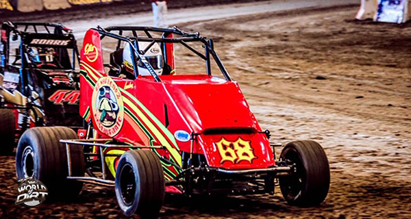 A red race car holds a driver with a white helmet as it drives on a dirt road with a black race car behind it.