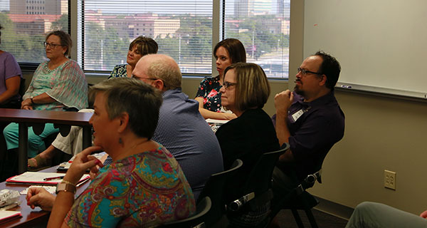 A group of men and women sit behind connected tables and watch something at the front of a room.