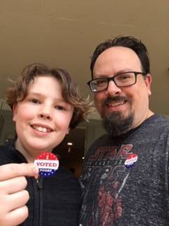 An older man faces the camera and stands to the left of his young son as both smile and the man wear an "I voted" pin and the boy hold the same pin in his right hand.