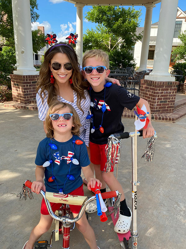 A woman hugging two children, all wearing clothes and holding pinwheels with american flags and red, white, and blue.