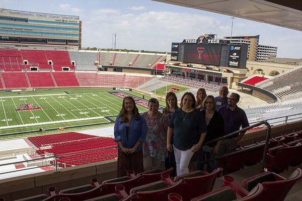 A group of men and women stand alongside one another while smiling and facing the camera with the football field of the Jones AT&T stadium in the background.