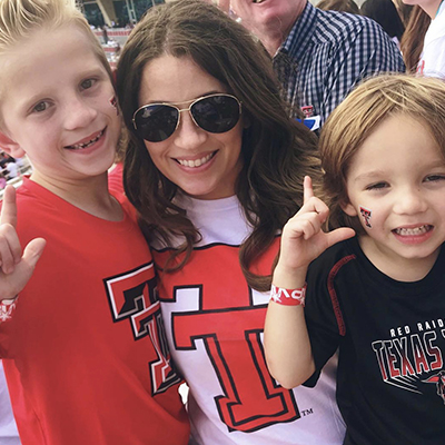 A woman wearing a white shirt with the Texas Tech logo is centered between two children that are displaying the guns up symbol while smiling.