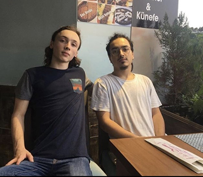 Two young men sit in chairs in a room behind a desk as they both look towards the camera