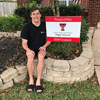 A young man sits outside around plants and a Texas Tech High School yard sign wearing a black shirt and sandles.