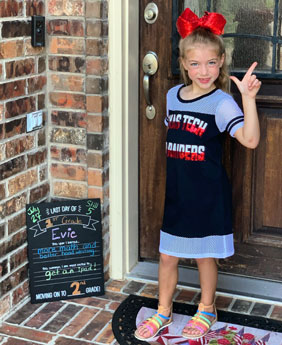 A young girl stands at the front of a residential door with a large red bow in her hair as she poses and flashes the guns up symbol and smiles