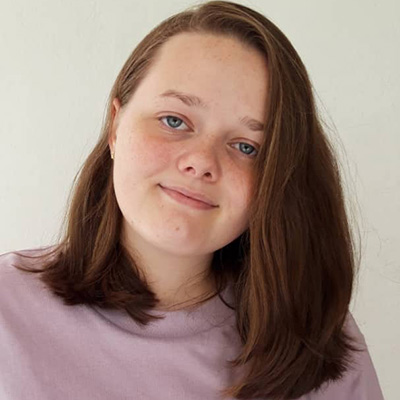 A young girl with brown hair looks at the camera while wearing a lavender colored shirt.