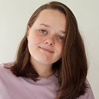 A young girl with brown hair looks at the camera while wearing a lavender colored shirt.