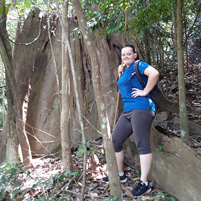 A young girl poses outdoors while standing next to trees wearing a blue shirt