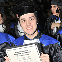 A young man wears a black graduation cap and a black gown and holds his high school diploma in his hands facing the camera.