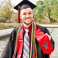 A young man wearing a black cap and gown and a red stole poses outdoors.