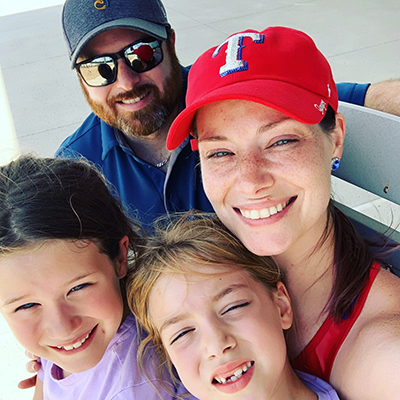 A man wearing sunglasses crouches behind a woman wearing a red Texas Rangers cap as she holds two small girls who squint at the camera.