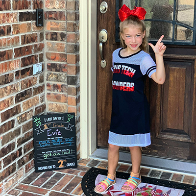 A young girl stands at the front of a residential door with a large red bow in her hair as she poses and flashes the guns up symbol and smiles