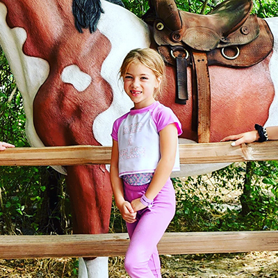 A young girl stands next to the body of fake horse as she smiles.