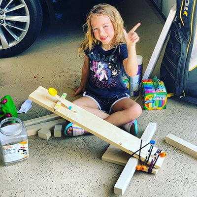 A young girl sits next to a car in a garage wearing a blue shirt with wooden planks in front of her while showing the guns up symbol.