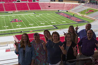 A group of men and women stand next to one another closely while some demonstrate the guns up symbol and others simply stand and smile with the Jones AT&T stadium field in the background