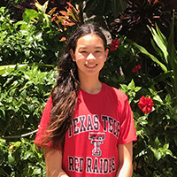 A young girl stands outdoors wearing a red Texas Tech shirt and smiles at the camera with red flowers in the background.