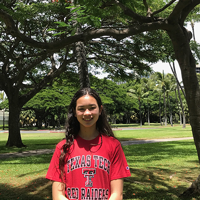 A young girl stands outdoors wearing a red Texas Tech shirt and smiles at the camera