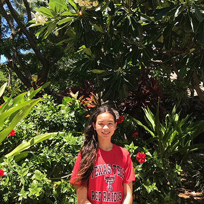 A young girl stands outdoors wearing a red Texas Tech shirt and smiles at the camera with red flowers in the background.