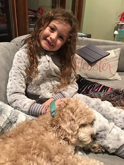 A young girl sits on a couch inside a home next to a small dog