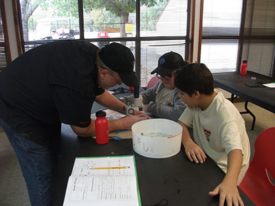 Mosley and his students study the water quality of the South Llano River, considering factors like pH, effects of temperature, and the life that call the river home. An older man leans over a desk with two students on the opposite end as they all look down at a project on the table.