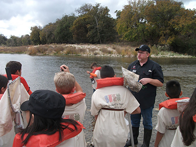 Mosley helps students identify macroinvertebrates with samples collected from the South Llano river. An older man stands in front of a group of kids wearing life preservers
