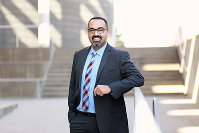A man wearing a gray suit and glasses stands outside with steps behind him as he smiles.