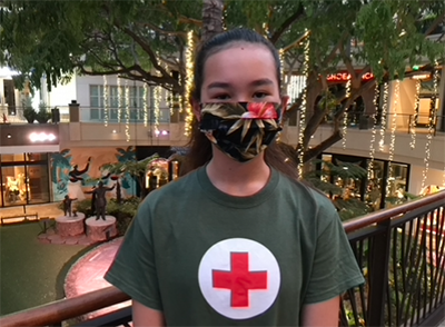 A young woman wearing a floral mask over her face and a Red Cross shirt stands next to railing indoors.