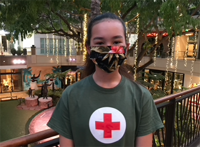 Kana is an American Red Cross National Outreach Coordinator. A young woman wearing a floral mask over her face and a Red Cross shirt stands next to railing indoors.