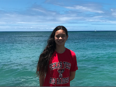 A young woman stands outdoors wearing a red Texas Tech shirt with a beautiful blue sky and ocean in the background.