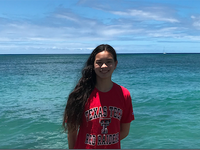 Kana Walsh shares the beauty of her home state. A young woman stands outdoors wearing a red Texas Tech shirt with a beautiful blue sky and ocean in the background.
