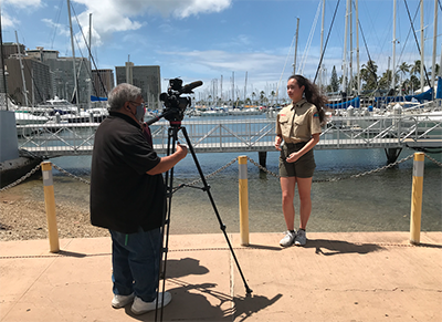 A man stands behind a video camera outdoors and records a young woman in front of him as both stands near water on a pier.