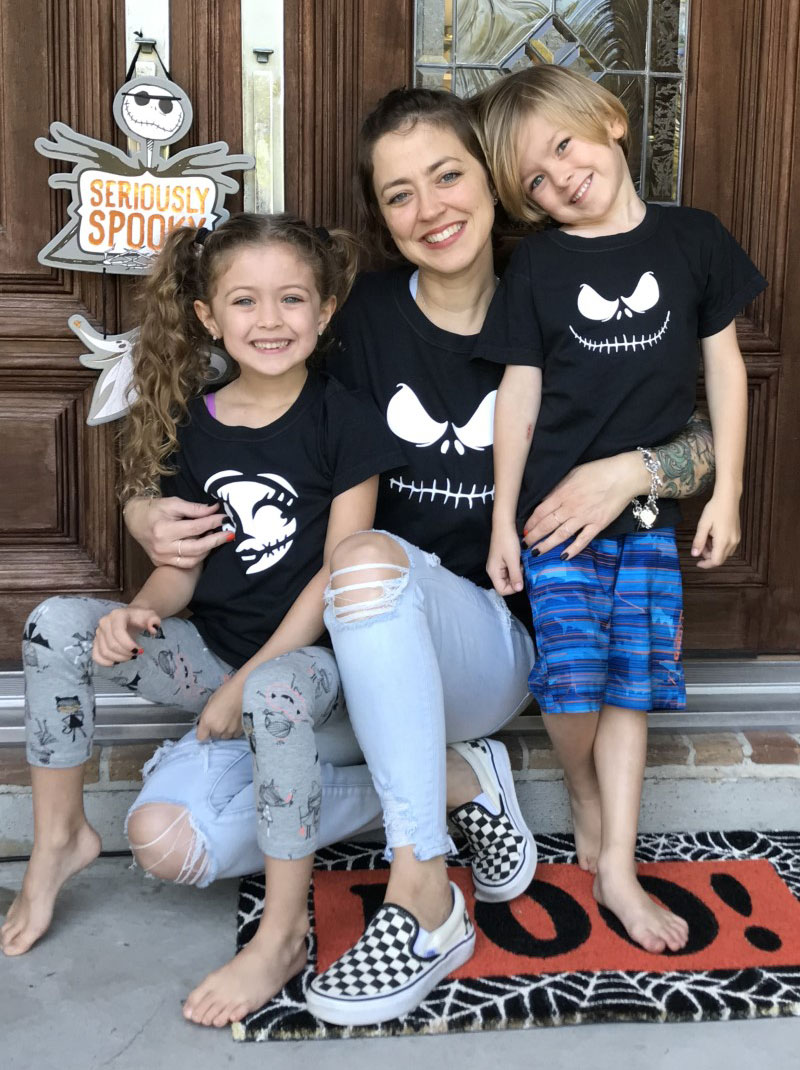 Kattie Brewer crouches between her two small children with the girl to the woman's right and the boy to the woman's left as all three wear black Nightmare Before Christmas shirts