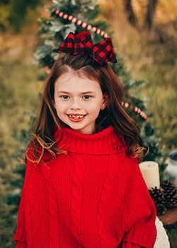A young girl wearing a red sweater and a red and black bow stands outdoors while smiling.
