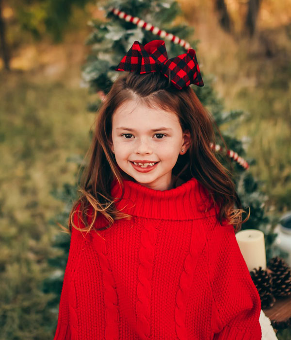 A young girl wearing a red sweater and a red and black bow stands outdoors while smiling.