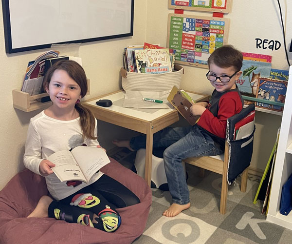 A young girl sits near her young brother as both face the camera smiling inside a room.