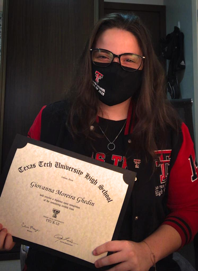 A woman wearing glasses and a Texas Tech mask holds her diploma in front of her body.