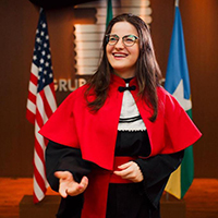 A young woman wearing glasses and a red, white, and black outfit stands inside a room with the American flag and another flag in the background as she smiles and looks off camera.