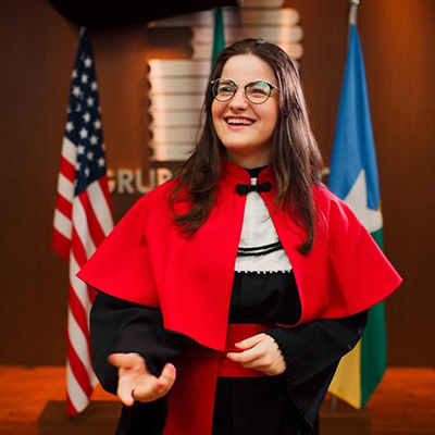 Giovanna is ready for commencement. A young woman wearing glasses and a red, white, and black outfit stands inside a room with the American flag and another flag in the background as she smiles and looks off camera.