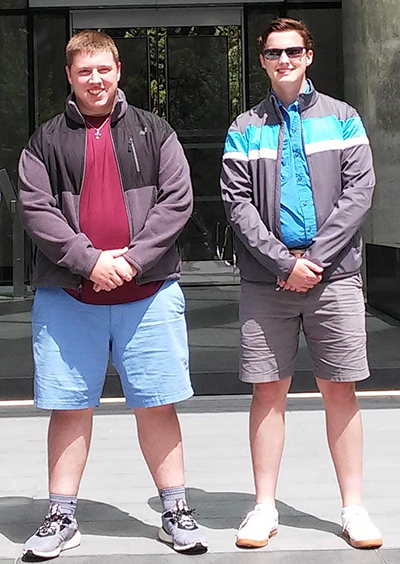 Two young men stand next to one another outdoors wearing coats and short pants while smiling.