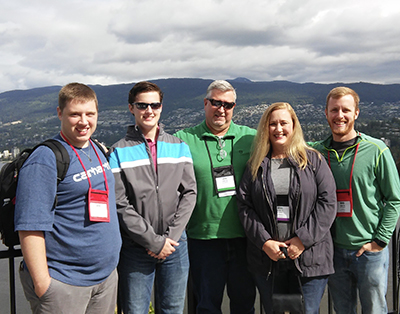 Three men and a woman all stand together outdoors with a scenic view behind them as they all smile while wearing lanyards around their necks.