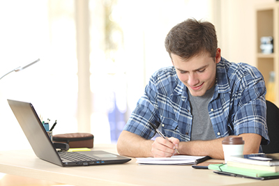 Student studying at desk A young man sits at a desk while wearing a blue shirt and writing something down on paper with a pen.