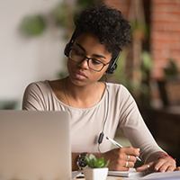 A young woman wearing glasses sits at a table where a laptop rests as she writes something on paper using a pen..