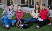 A group of students consisting of a young male and three females all sit outdoors while smiling and performing the guns up symbol.