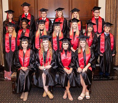 A large group of young men and women stand together wearing red stoles, black graduation caps, and black gowns while smiling at the camera.
