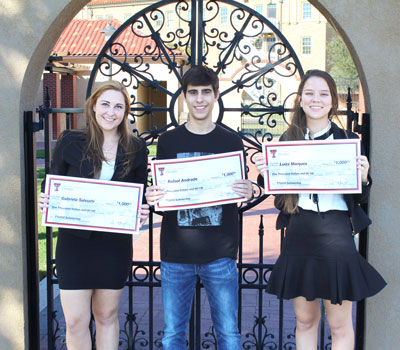 A young man stands between two young women as all three hold large checks in their hands while standing outdoors and smiling.