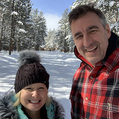 A shorter woman and a taller male stand next to one another with snow covered trees in the background and a large sheet of snow on the ground.