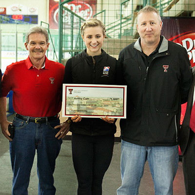 Two men stand on either side of a young woman as she holds a large check in her hands.