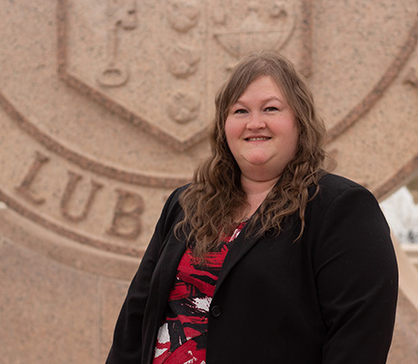 A woman wearing a black blazer and red shirt stands outdoors in front of the Texas Tech University seal.