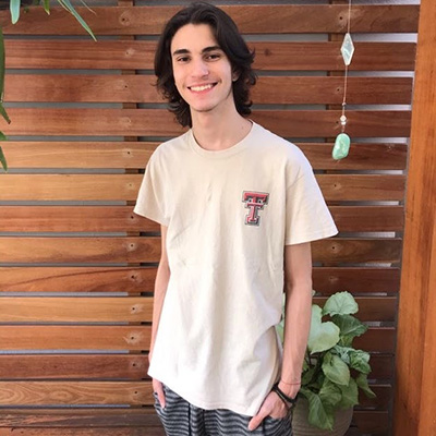 A young man wearing a white Texas Tech shirt smiles as he stands in front of a wooden fixture.