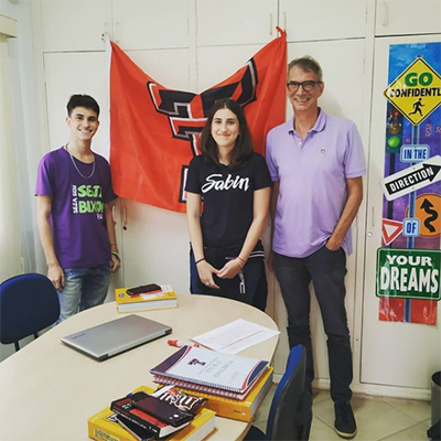 A young man stands alongside a man and woman who all stand in front of a Texas Tech flag and behind a table with a laptop and books resting on top.